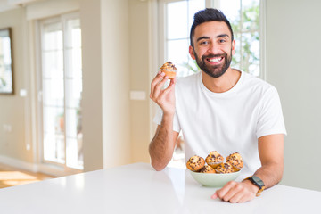 Handsome hispanic man eating chocolate chips muffin with a happy face standing and smiling with a...