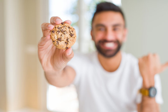 Handsome hispanic man eating chocolate chips cookies pointing and showing with thumb up to the side with happy face smiling