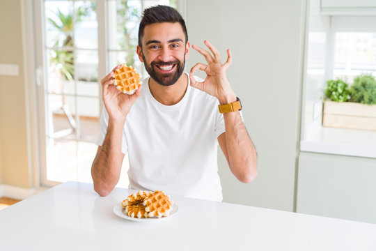 Handsome hispanic man eating sweet belgian waffle pastry doing ok sign with fingers, excellent symbol