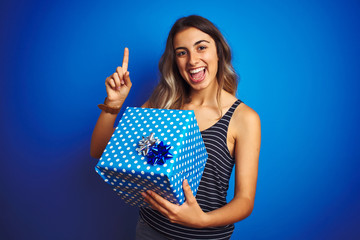 Young woman holding birthday present over blue isolated background surprised with an idea or question pointing finger with happy face, number one