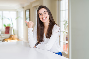 Beautiful young woman sitting on white table at home smiling with happy face looking and pointing to the side with thumb up.