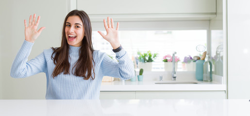 Wide angle picture of beautiful young woman sitting on white table at home showing and pointing up with fingers number ten while smiling confident and happy.