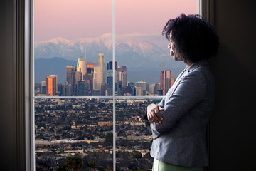 Black female business woman looking out the window of an office in Los Angeles.  She looks like a...