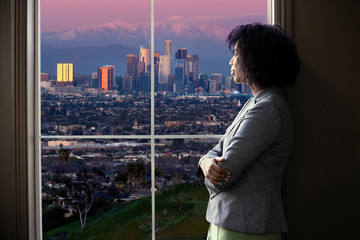 Black female business woman looking out the window of an office in Los Angeles.  She looks like a...