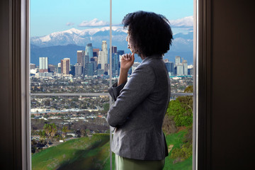 Black female business woman looking out the window of an office in Los Angeles.  She looks like a...