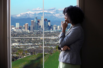 Black female business woman looking out the window of an office in Los Angeles.  She looks like a...