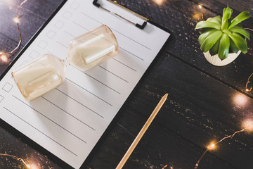 clipboard with blank list with cases to tick off on wooden desk with hourglass