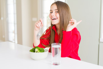 Beautiful young girl kid eating fresh broccoli and drinking water very happy pointing with hand and finger to the side