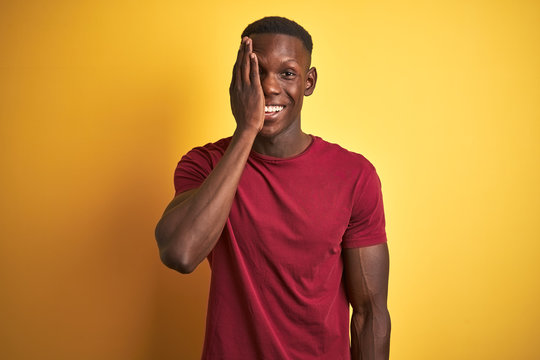 Young african american man wearing red t-shirt standing over isolated yellow background covering one eye with hand, confident smile on face and surprise emotion.