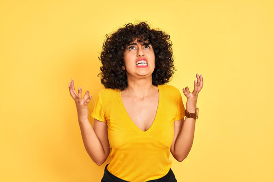 Young arab woman with curly hair wearing t-shirt standing over isolated yellow background crazy and mad shouting and yelling with aggressive expression and arms raised. Frustration concept.