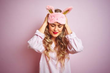 Young beautiful woman wearing pajama and sleep mask over pink isolated background suffering from headache desperate and stressed because pain and migraine. Hands on head.