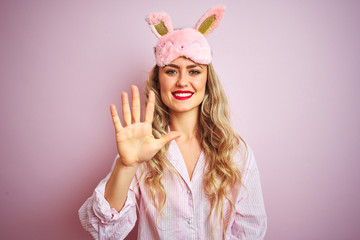 Obraz na płótnie Canvas Young beautiful woman wearing pajama and sleep mask over pink isolated background showing and pointing up with fingers number five while smiling confident and happy.