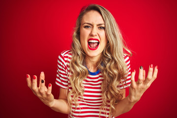 Young beautiful woman wearing stripes t-shirt standing over red isolated background crazy and mad shouting and yelling with aggressive expression and arms raised. Frustration concept.