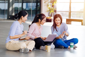 Group of young smart Asian students laughing making notes looking at laptop and discussing while sitting on floor outdoors