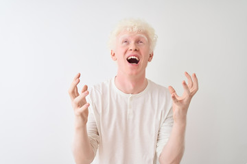 Young albino blond man wearing casual t-shirt standing over isolated white background crazy and mad shouting and yelling with aggressive expression and arms raised. Frustration concept.