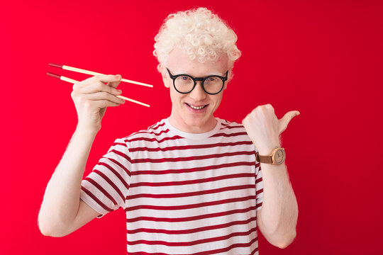 Young albino blond man holding chopsticks standing over isolated white background pointing and showing with thumb up to the side with happy face smiling