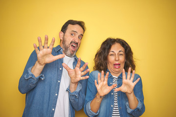 Beautiful middle age couple together wearing denim shirt over isolated yellow background afraid and terrified with fear expression stop gesture with hands, shouting in shock. Panic concept.