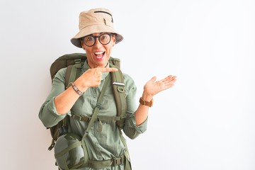 Middle age hiker woman wearing backpack canteen hat glasses over isolated white background amazed and smiling to the camera while presenting with hand and pointing with finger.