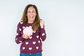 Middle age woman holding piggy bank over isolated background screaming proud and celebrating victory and success very excited, cheering emotion