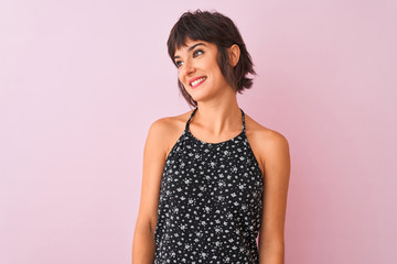 Young beautiful woman wearing black floral dress standing over isolated pink background looking away to side with smile on face, natural expression. Laughing confident.