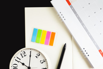 Close up pen, clock, sticky post and calendar placed on blank notebook with black desk background in dramatic lighting tone. Concept for business, planning or diary.Great for any use.Clipping Path.