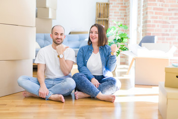 Young couple sitting on the floor arround cardboard boxes moving to a new house smiling with happy face looking and pointing to the side with thumb up.