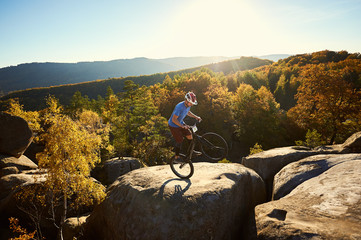 Fototapeta na wymiar Young cyclist standing on back wheel on trial bicycle, sportsman rider making acrobatic trick on the edge of big boulder on the top of mountain at sunset. Concept of extreme sport active lifestyle