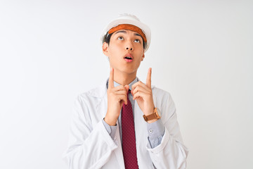 Chinese architect man wearing coat and helmet standing over isolated white background amazed and surprised looking up and pointing with fingers and raised arms.
