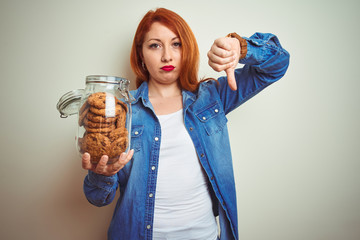 Young beautiful redhead woman holding jar of cookies over white isolated background with angry...