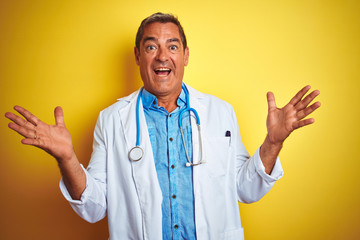 Handsome middle age doctor man wearing stethoscope over isolated yellow background celebrating crazy and amazed for success with arms raised and open eyes screaming excited. Winner concept