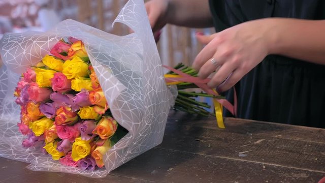 Floral business concept. Woman florist is packaging bouquet of small colourful roses in paper in flower shop and ties ribbon on stems, hands closeup. She works in professiona floristic studio, store.
