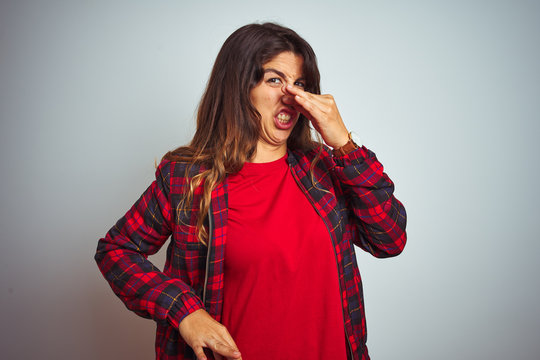 Young beautiful woman wearing red t-shirt and jacket standing over white isolated background smelling something stinky and disgusting, intolerable smell, holding breath with fingers on nose. 