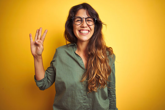 Young beautiful woman wearing green shirt and glasses over yelllow isolated background showing and pointing up with fingers number four while smiling confident and happy.