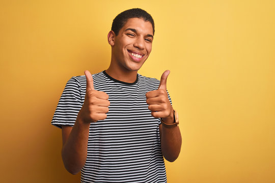 Young handsome arab man wearing navy striped t-shirt over isolated yellow background success sign doing positive gesture with hand, thumbs up smiling and happy. Cheerful expression and winner gesture.