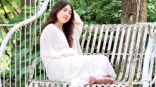 Beautiful Chinese young woman in white dress and barefoot sitting on swing chair swing to sleep, beauty in summer, 4k footage, slow motion.