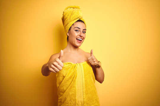 Young beautiful woman wearing towel after shower over isolated yellow background approving doing positive gesture with hand, thumbs up smiling and happy for success. Winner gesture.