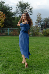 A young woman in a blue dress walks barefoot on the grass in the park