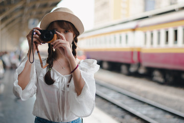 Pretty Asian girl wearing a white shirt and hat, standing at the train station, in her hands holding a camera, she is a tourist who likes to take pictures.