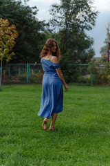 A young woman in a blue dress walks barefoot on the grass in the park