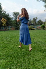 A young woman in a blue dress walks barefoot on the grass in the park