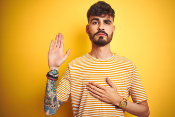 Young man with tattoo wearing striped t-shirt standing over isolated yellow background Swearing with hand on chest and open palm, making a loyalty promise oath