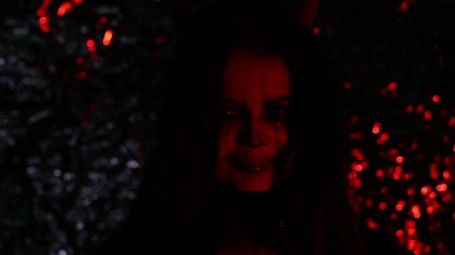 Girl in the form of a devil with horns. Image of halloween in red light. Contact lenses and a scary look.