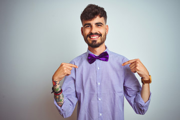 Young man with tattoo wearing purple shirt and bow tie over isolated white background looking...
