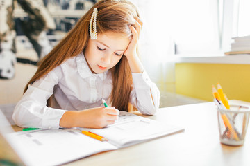Education at home concept - Cute little girl with long hair studying or completing home work on a table with pile of books and workbook