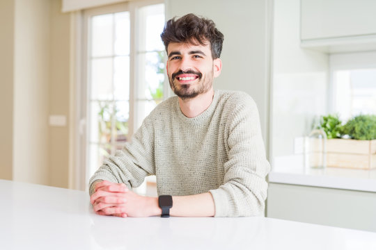 Young man wearing casual sweater sitting on white table with a happy and cool smile on face. Lucky person.