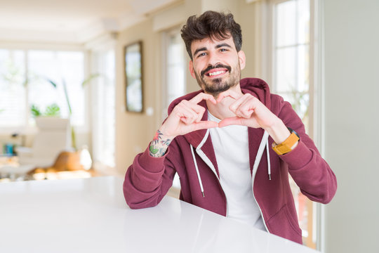 Young man wearing casual sweatshirt sitting on white table smiling in love showing heart symbol and shape with hands. Romantic concept.