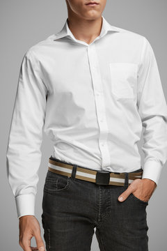 Cropped medium full shot of a young man dressed in a white buttoned shirt with a pocket and gray jeans with a belt. The goldish belt with a white stripe is made of textured fabric and equipped