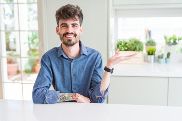 Young man wearing casual shirt sitting on white table smiling cheerful presenting and pointing with palm of hand looking at the camera.