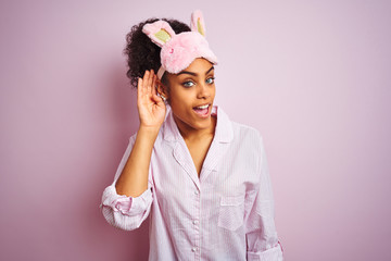 Fototapeta na wymiar Young african american woman wearing pajama and mask over isolated pink background smiling with hand over ear listening an hearing to rumor or gossip. Deafness concept.