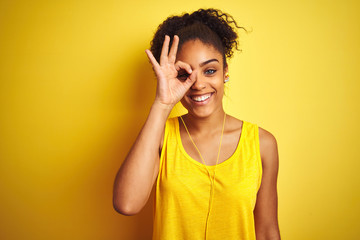 African american woman listening to music using earphones over isolated yellow background doing ok gesture with hand smiling, eye looking through fingers with happy face.